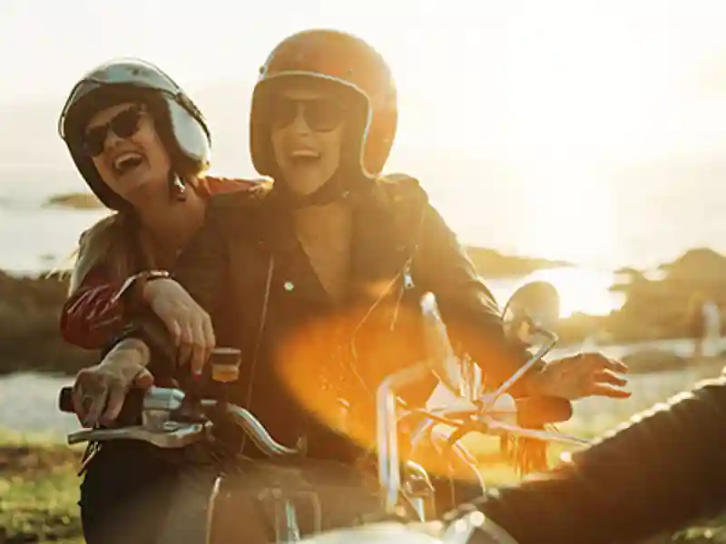 Two women laugh wholeheartedly as they prepare to ride a motorcycle together.