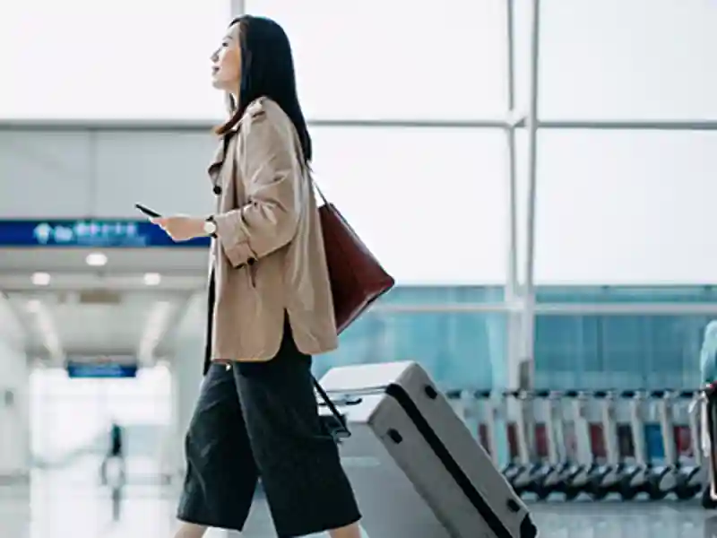 A woman walks through an airport with her passport and roller bag.