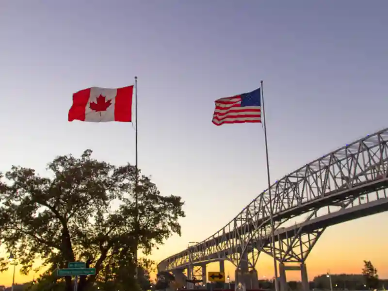 Canada and U.S. flags wave in front of a bridge at sunset.