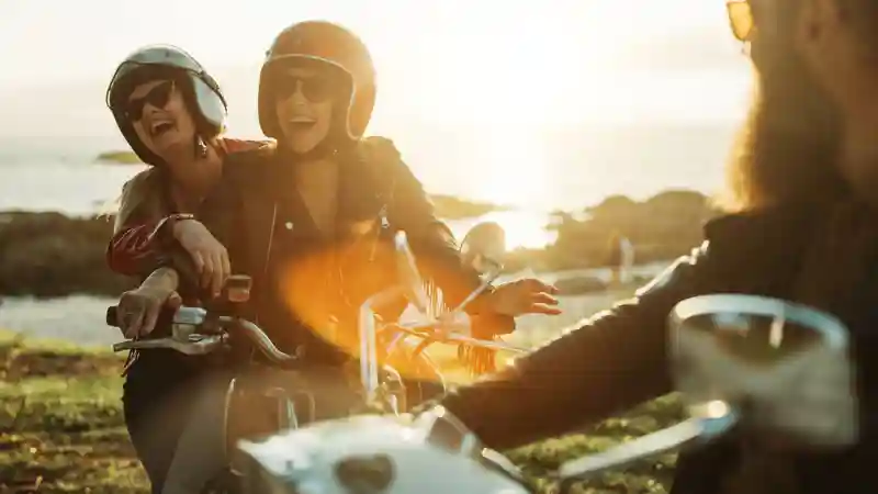Two women wearing helmets and sunglasses ride a motorcycle, showcasing a sense of adventure while laughing.