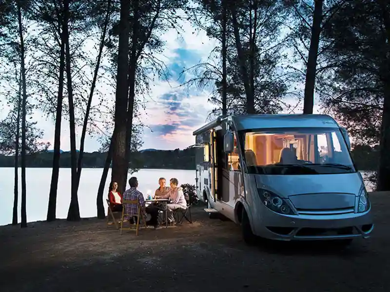 A group of people converse at a lakeside campground next to a large recreational vehicle.