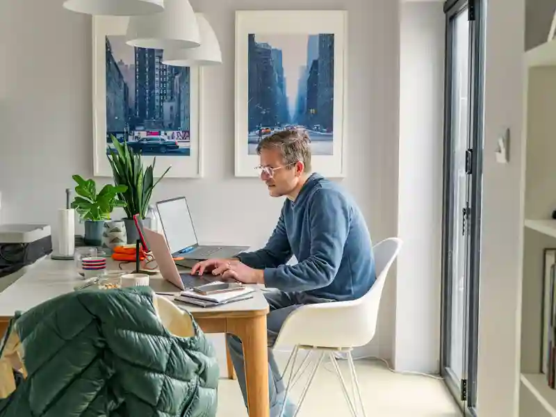 A casually-dressed man works on two laptops in a home office